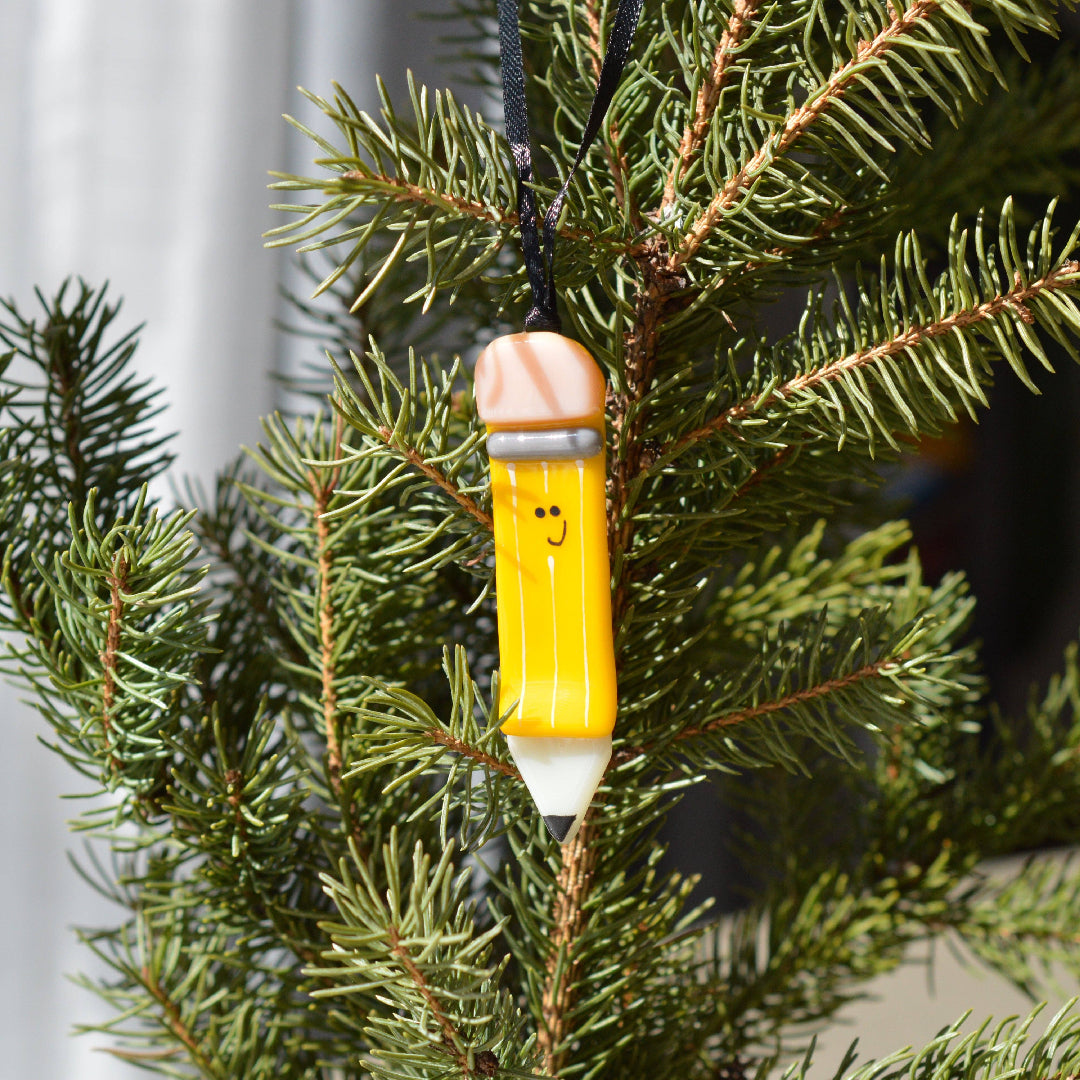 A yellow pencil-shaped Christmas ornament with a smiling face hangs from a black ribbon among green pine branches.