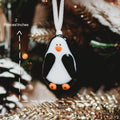 A penguin-shaped glass ornament hanging from a white ribbon, with overlaid dimension arrows and text showing 2 pouces/10 cm height and 1 1/2 pouces/6 cm width, against a blurred Christmas tree background.