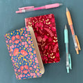 Two floral notebooks with pens on a teal background