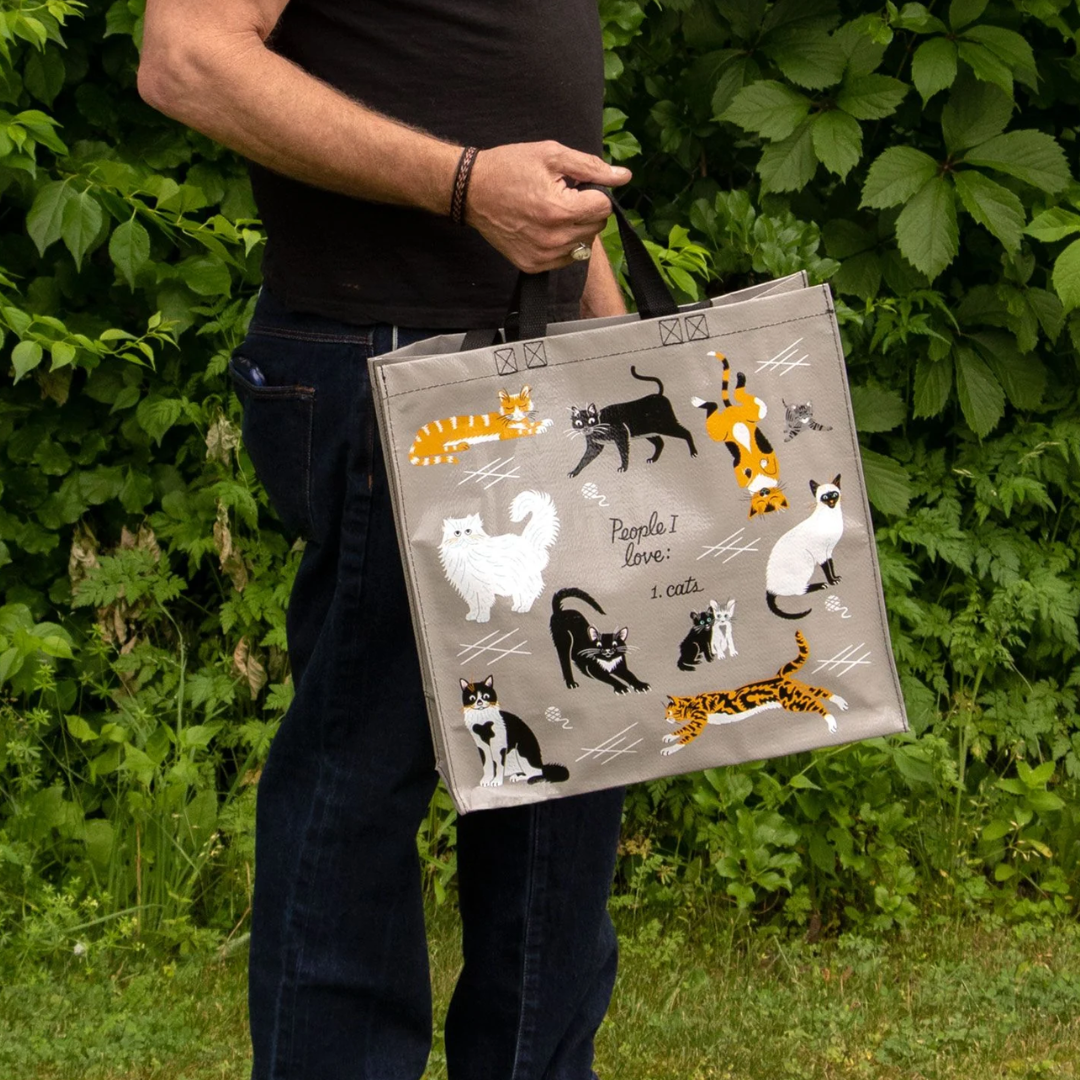 A reusable tote bag with a cat-themed design. The bag is a light grey color with black handles. It features a variety of cats in different poses and breeds, including a white fluffy cat, a black cat, a tabby cat, and a Siamese cat. The text "People I love: 1. Cats" is printed on the bag in black.