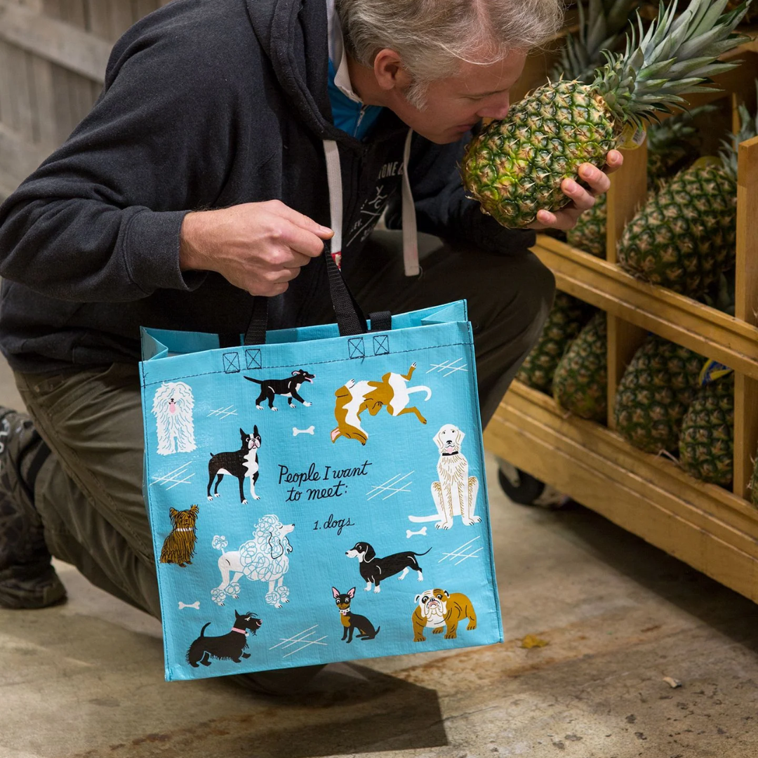 A reusable tote bag with a playful dog-themed design. The bag is a light blue color with black handles. It features a variety of dog breeds in different poses, including a bulldog, a poodle, a dachshund, and a husky. The text "People I want to meet: 1. Dogs" is printed on the bag in black.