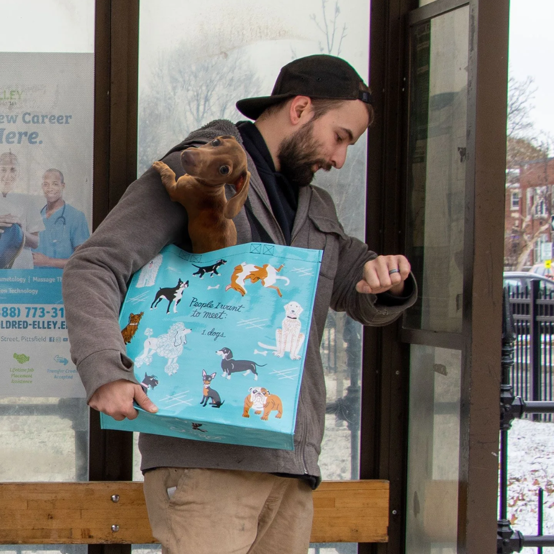 A reusable tote bag with a playful dog-themed design. The bag is a light blue color with black handles. It features a variety of dog breeds in different poses, including a bulldog, a poodle, a dachshund, and a husky. The text "People I want to meet: 1. Dogs" is printed on the bag in black.