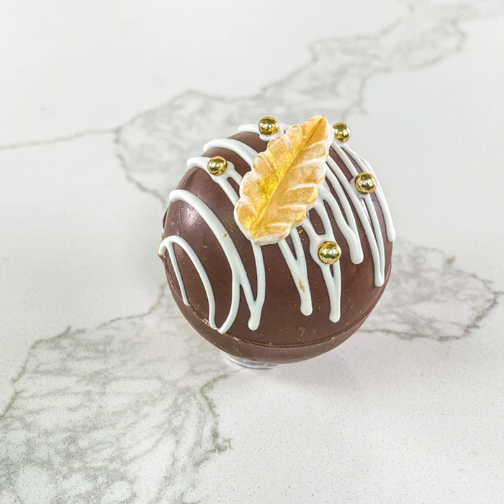 A dark chocolate hot chocolate bomb decorated with white drizzle, a gold leaf, and gold beads. The bomb is resting on a white marble surface.