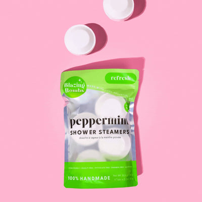 A vibrant, high-angle flat lay shot on a pink background features a shiny, white and bright green pouch of Blazing Bombs Peppermint Shower Steamers. The pouch is labeled 