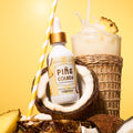 A bottle of Ananda do Brasil Piña Colada (pineapple cologne) styled with a coconut, pineapple slices, a straw, and a tropical drink on a yellow background.