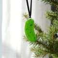A bright green pickle-shaped glass ornament hanging from a black ribbon, positioned next to green pine tree branches against a soft white curtain background.