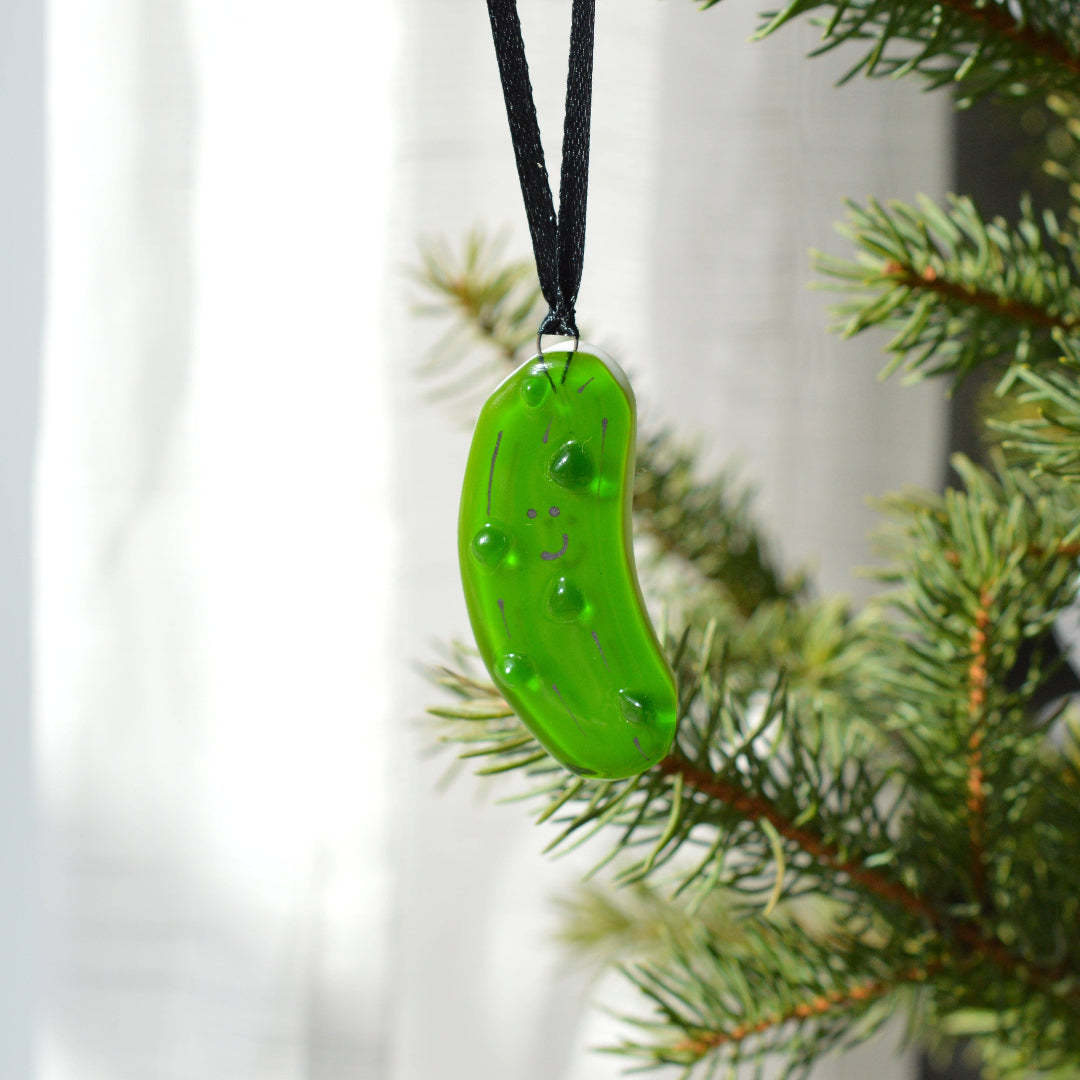 A bright green pickle-shaped glass ornament hanging from a black ribbon, positioned next to green pine tree branches against a soft white curtain background.