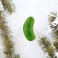 A green pickle-shaped Christmas ornament with a smiling face, hanging from a white ribbon, surrounded by pine branches and fairy lights on a white background.