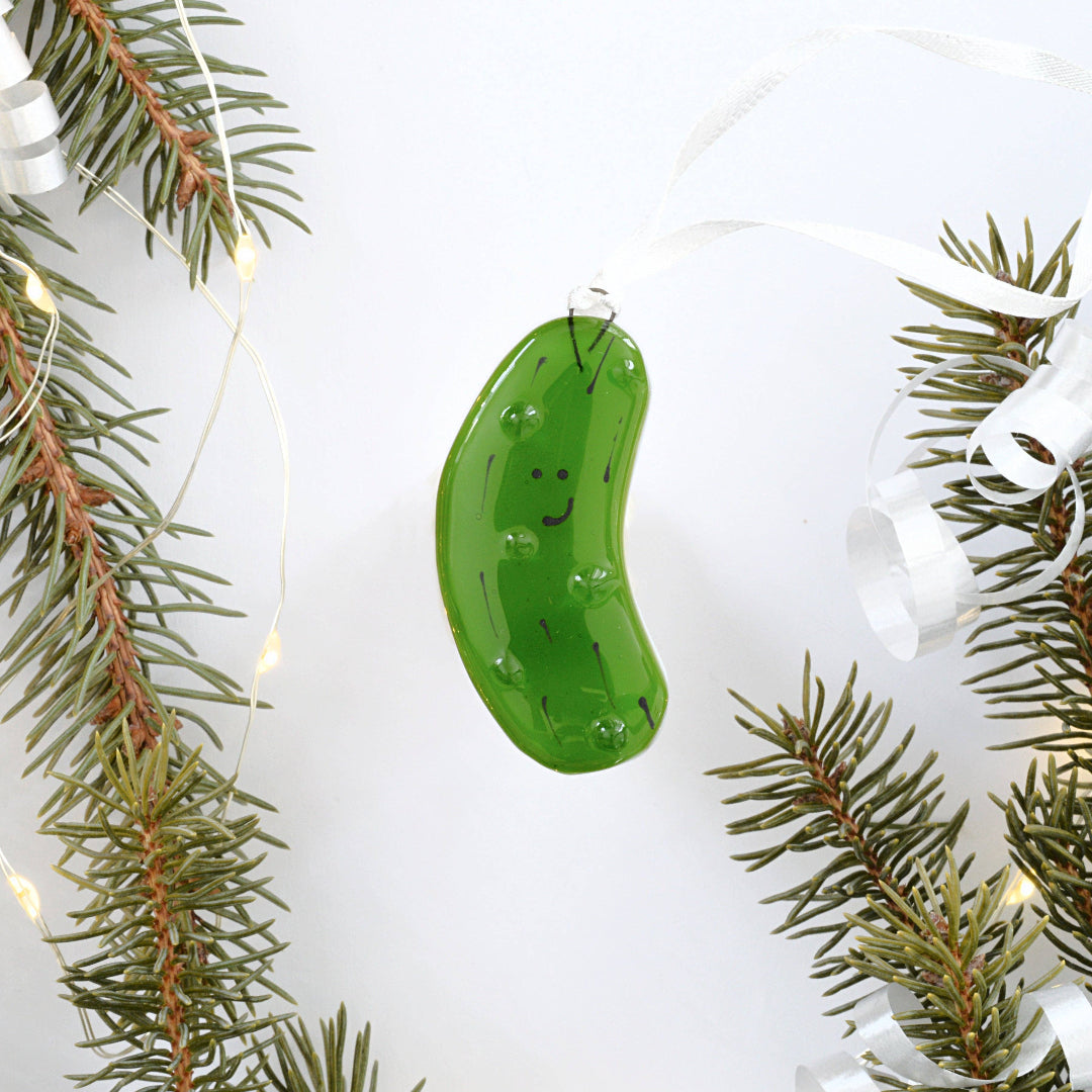 A green pickle-shaped Christmas ornament with a smiling face, hanging from a white ribbon, surrounded by pine branches and fairy lights on a white background.