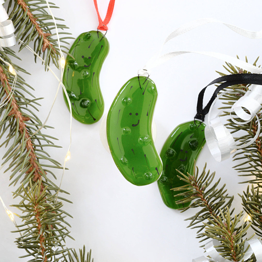 Three green pickle-shaped glass ornaments with small black facial features and water droplets, hanging from red, silver, and black ribbons and styled with pine branches and fairy lights on a white background.