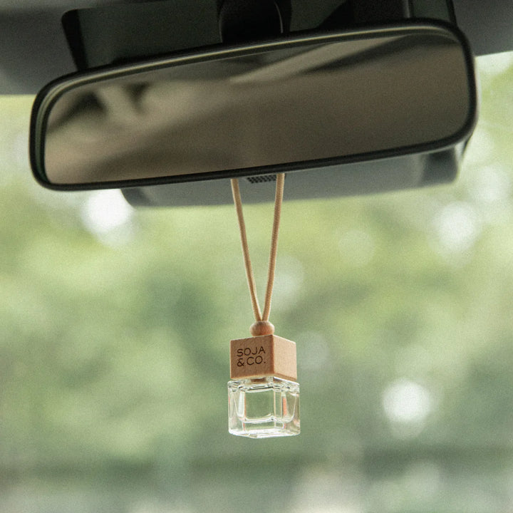 A car diffuser kit by Soja & Co. The kit includes a small glass diffuser bottle with a wooden cap and a string to hang it from the rearview mirror. Next to it is a cardboard box with the brand logo and product information. The box is labeled 