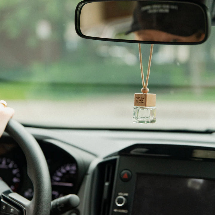 A car diffuser kit by Soja & Co. The kit includes a small glass diffuser bottle with a wooden cap and a string to hang it from the rearview mirror. Next to it is a cardboard box with the brand logo and product information. The box is labeled 