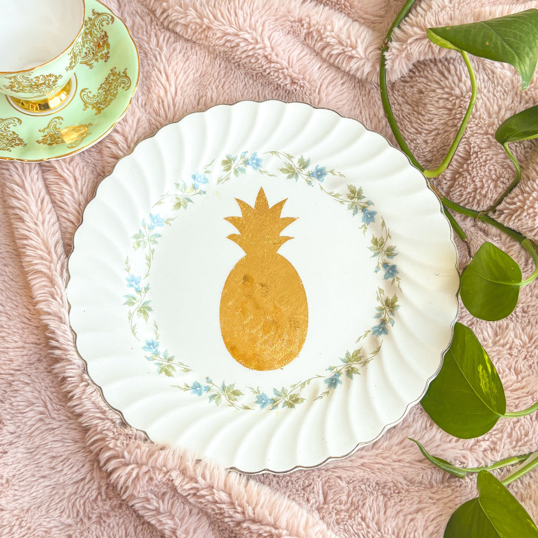 White plate with gold pineapple design on a pink textured surface with green leaves.