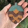 A hand holds a felt pinecone-shaped ornament or decoration with layered green, tan, and brown scales, set against evergreen branches.