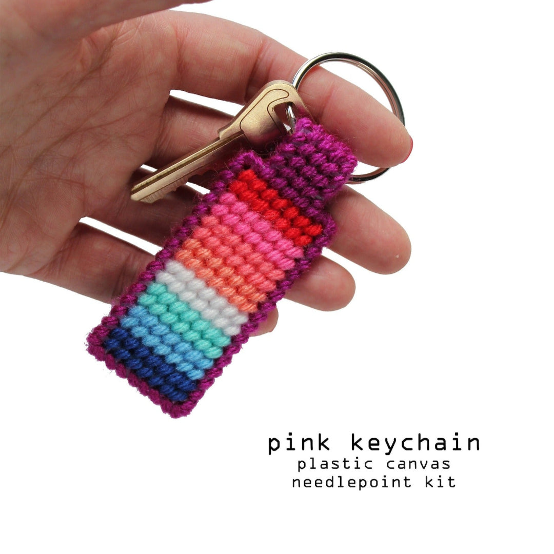 A hand holds a finished needlepoint keychain and a set of keys. The keychain is rectangular with a striped pattern in shades of pink, orange, yellow, green, and blue, and is attached to a silver key ring.