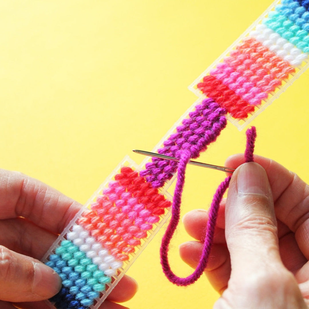 A hand holds a finished needlepoint keychain and a set of keys. The keychain is rectangular with a striped pattern in shades of pink, orange, yellow, green, and blue, and is attached to a silver key ring.