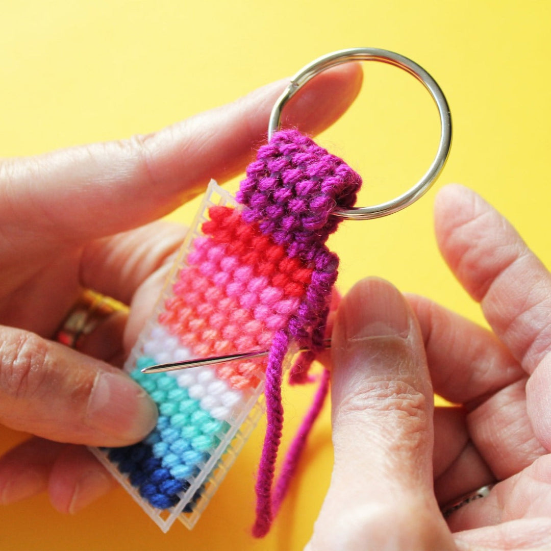 A hand holds a finished needlepoint keychain and a set of keys. The keychain is rectangular with a striped pattern in shades of pink, orange, yellow, green, and blue, and is attached to a silver key ring.