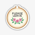A circular sticker designed to look like an embroidery hoop, holding a piece of cream-coloured fabric. Cross-stitched in black on the fabric are the words "PLEASE LEAVE". Below the text, there's a cross-stitched floral design with three pink flowers and green leaves.