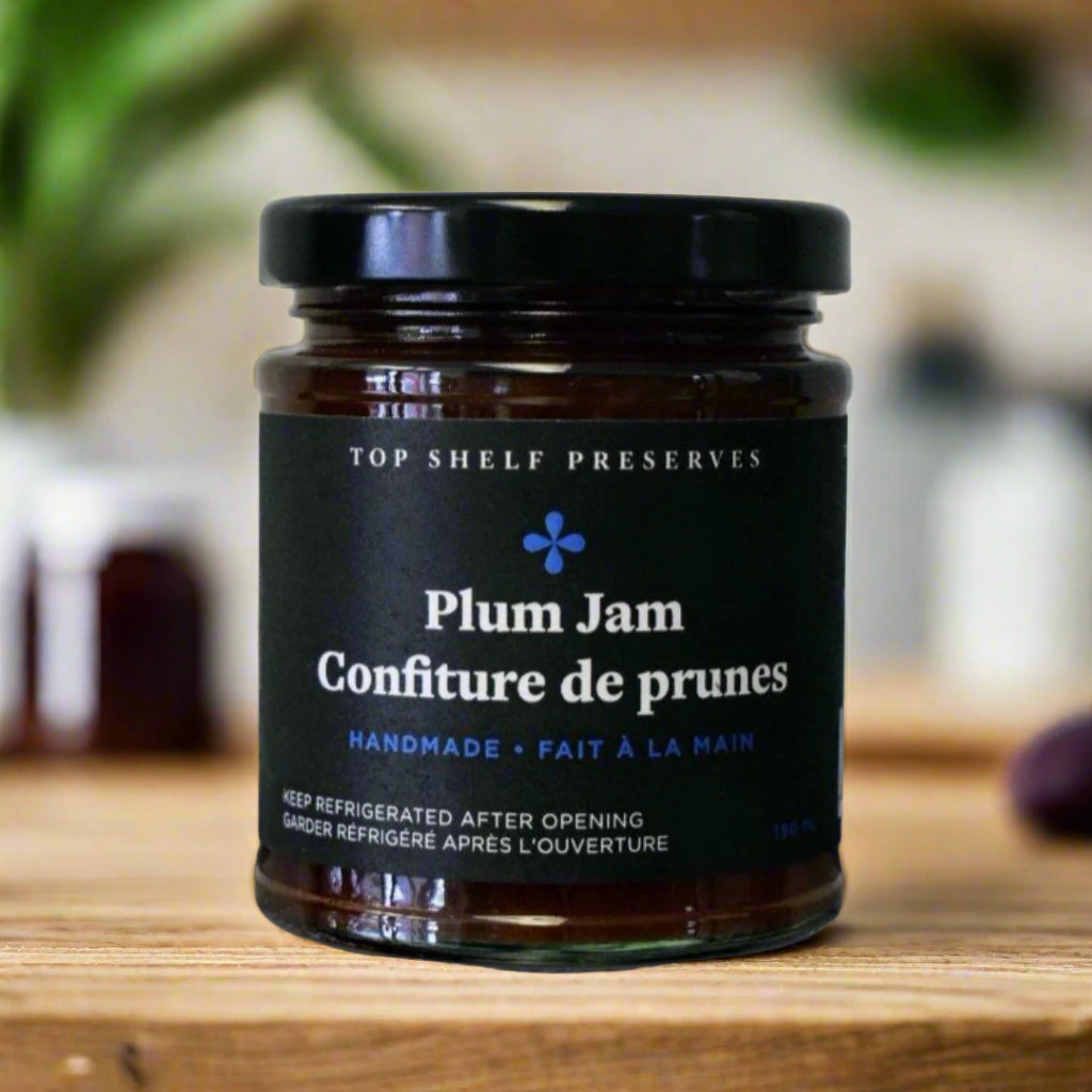 A jar of Plum Jam from Top Shelf Preserves. The jar is filled with a purple jam and has a black lid. The label is black with white text, including the product name, "Handmade Fait à la Main," and instructions to keep refrigerated after opening. 
