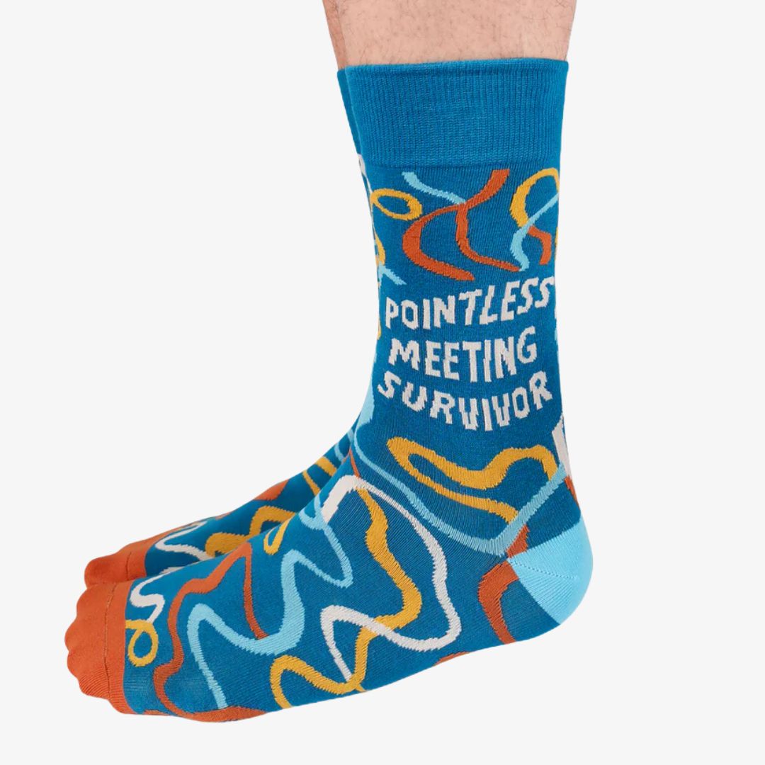 A pair of blue crew socks featuring a colourful confetti-like pattern in shades of orange, yellow, and white. One sock displays the text "POINTLESS MEETING SURVIVOR" in white letters. The toe of the sock is orange, the heel is light blue, and the cuff is a slightly darker blue. The brand "Uptown Sox" is subtly visible on the sole.