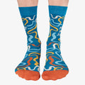 A pair of blue crew socks featuring a colourful confetti-like pattern in shades of orange, yellow, and white. One sock displays the text "POINTLESS MEETING SURVIVOR" in white letters. The toe of the sock is orange, the heel is light blue, and the cuff is a slightly darker blue. The brand "Uptown Sox" is subtly visible on the sole.