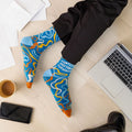 A pair of blue crew socks featuring a colourful confetti-like pattern in shades of orange, yellow, and white. One sock displays the text "POINTLESS MEETING SURVIVOR" in white letters. The toe of the sock is orange, the heel is light blue, and the cuff is a slightly darker blue. The brand "Uptown Sox" is subtly visible on the sole.