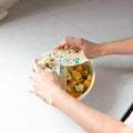 Person using a reusable food wrap on a bowl of fruit on a kitchen counter.