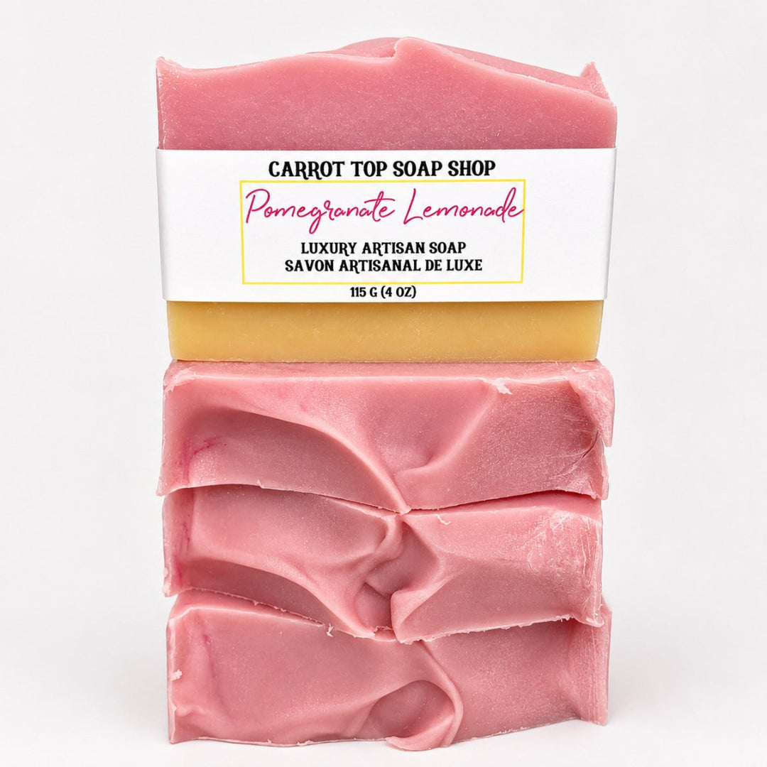 Three stacked bars of handmade artisan soap from Carrot Top Soap Shop, labeled 'Pomegranate Lemonade' in pink, with a white paper label and a yellow wax layer visible between the bars, on a white background.