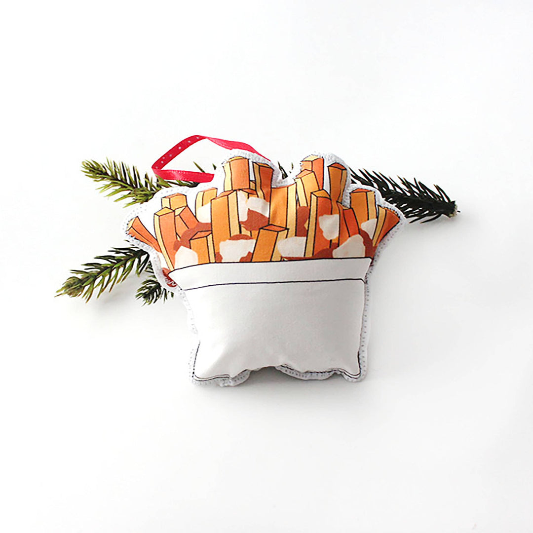 A close-up, high-angle flat lay shot on a plain white background features a soft, plush Christmas ornament shaped like a serving of poutine. The illustration shows a white cup filled with golden-brown french fries and a small amount of gravy or cheese curds. A red polka-dot ribbon loop is attached to the top for hanging.