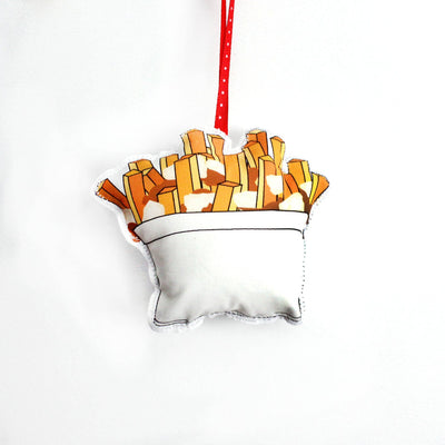 A close-up, high-angle flat lay shot on a plain white background features a soft, plush Christmas ornament shaped like a serving of poutine. The illustration shows a white cup filled with golden-brown french fries and a small amount of gravy or cheese curds. A red polka-dot ribbon loop is attached to the top for hanging.
