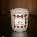 A North of Maine 'Pumpkin Cardamom' scented candle in a white glass jar, featuring a brown and white argyle pattern on the label, set on a dark wooden surface against a dark wood background.