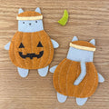 Two handmade felt cat plush toys dressed as pumpkins for Halloween, displayed on a light wood background. One cat is shown from the front wearing a jack-o'-lantern face, and the other is shown from the back with a stitched tail.