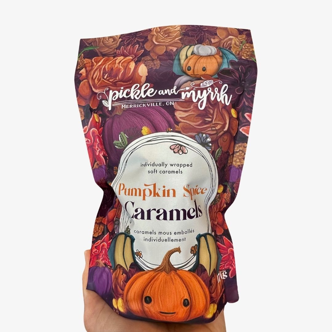 A hand holding a bag of Pickle and Myrk Pumpkin Spice Caramels against a plain white background. The packaging features a colorful autumn pattern with pumpkins, fall leaves, and a small cartoon character.