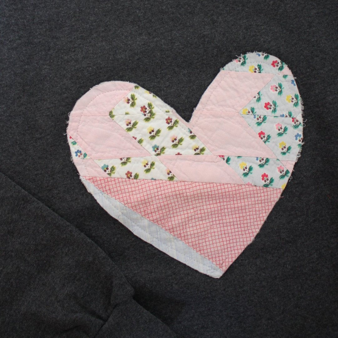 a grey sweatshirt with a large, heart-shaped patch. The patch is made from various fabrics with various patterns, sewn together in a patchwork style.