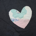 a grey sweatshirt with a large, heart-shaped patch. The patch is made from various fabrics with various patterns, sewn together in a patchwork style.