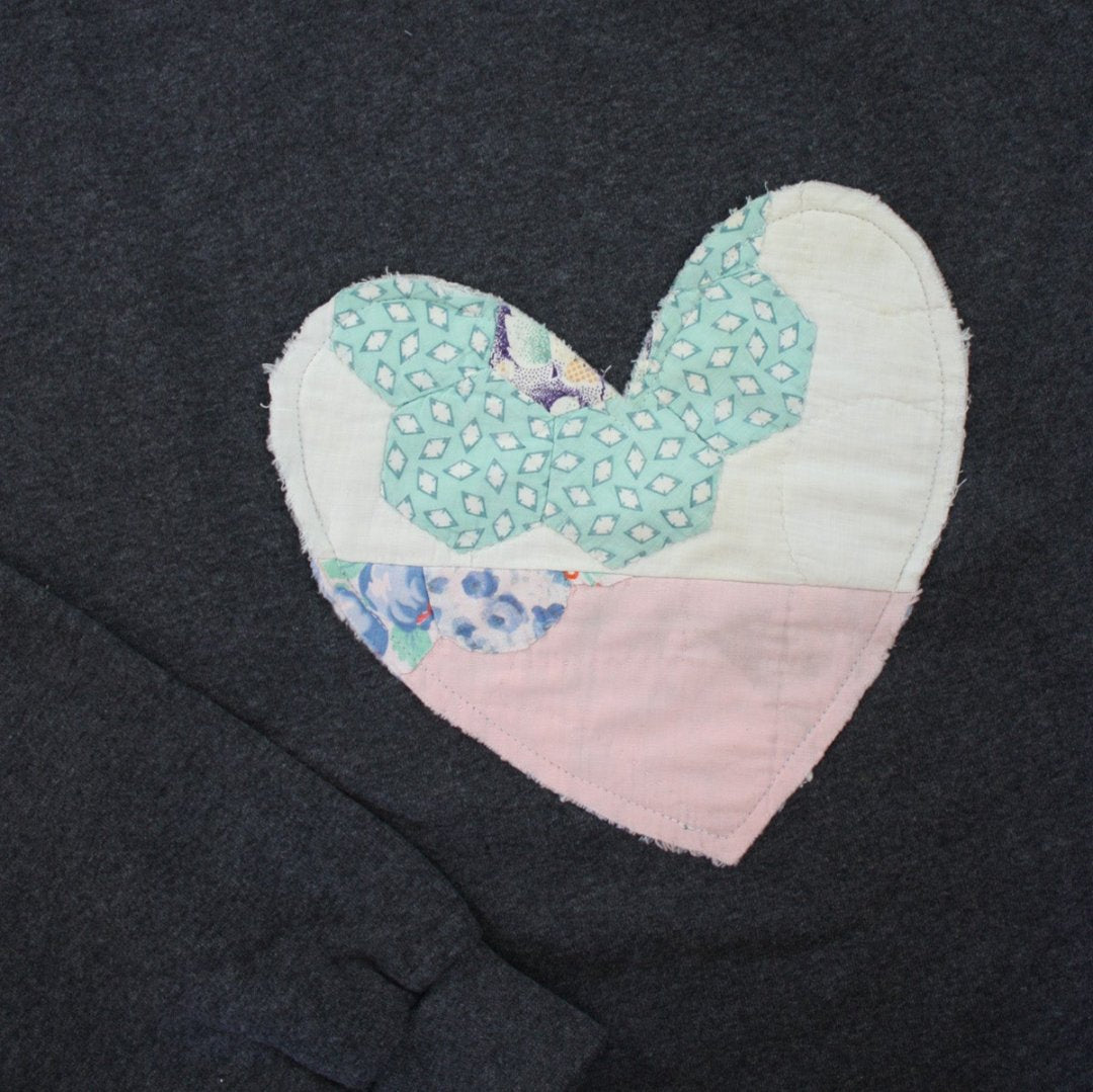 a grey sweatshirt with a large, heart-shaped patch. The patch is made from various fabrics with various patterns, sewn together in a patchwork style.