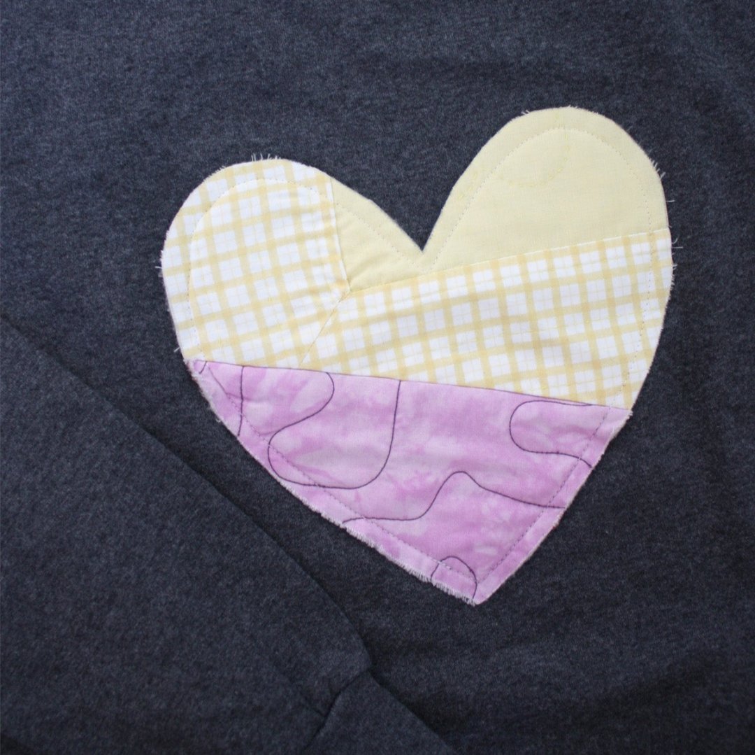 a grey sweatshirt with a large, heart-shaped patch. The patch is made from various fabrics with various patterns, sewn together in a patchwork style.