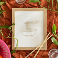Framed artwork of a bowl with chopsticks on a rust-colored fabric background