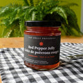 A jar of Red Pepper Jelly from Top Shelf Preserves. The jar is filled with a red jelly and has a black label with white text. The label includes the product name, "Handmade Fait à la Main," and instructions to keep refrigerated after opening.