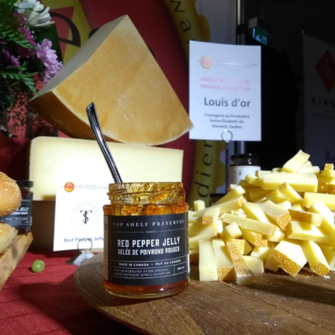 A jar of Red Pepper Jelly from Top Shelf Preserves. The jar is filled with a red jelly and has a black label with white text. The label includes the product name, "Handmade Fait à la Main," and instructions to keep refrigerated after opening.