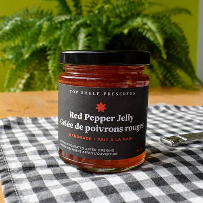 A jar of Red Pepper Jelly from Top Shelf Preserves. The jar is filled with a red jelly and has a black label with white text. The label includes the product name, 