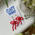 Red bow-shaped earrings on a 'CAIT WIND' branded card with plants in the background.