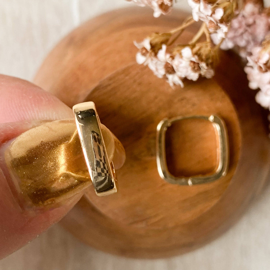 Gold hoop earrings held by a hand with a wooden coaster and flowers in the background