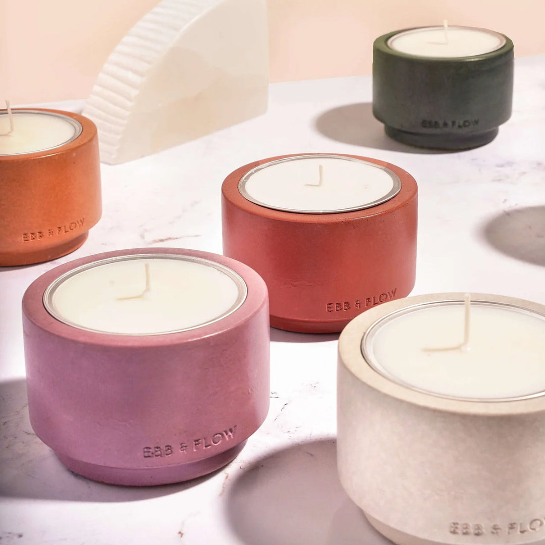 Four matte ceramic tea light candles in different earthy colors (terracotta, rust red, dusty mauve, and textured cream) arranged on a light marble surface with a soft beige background.
