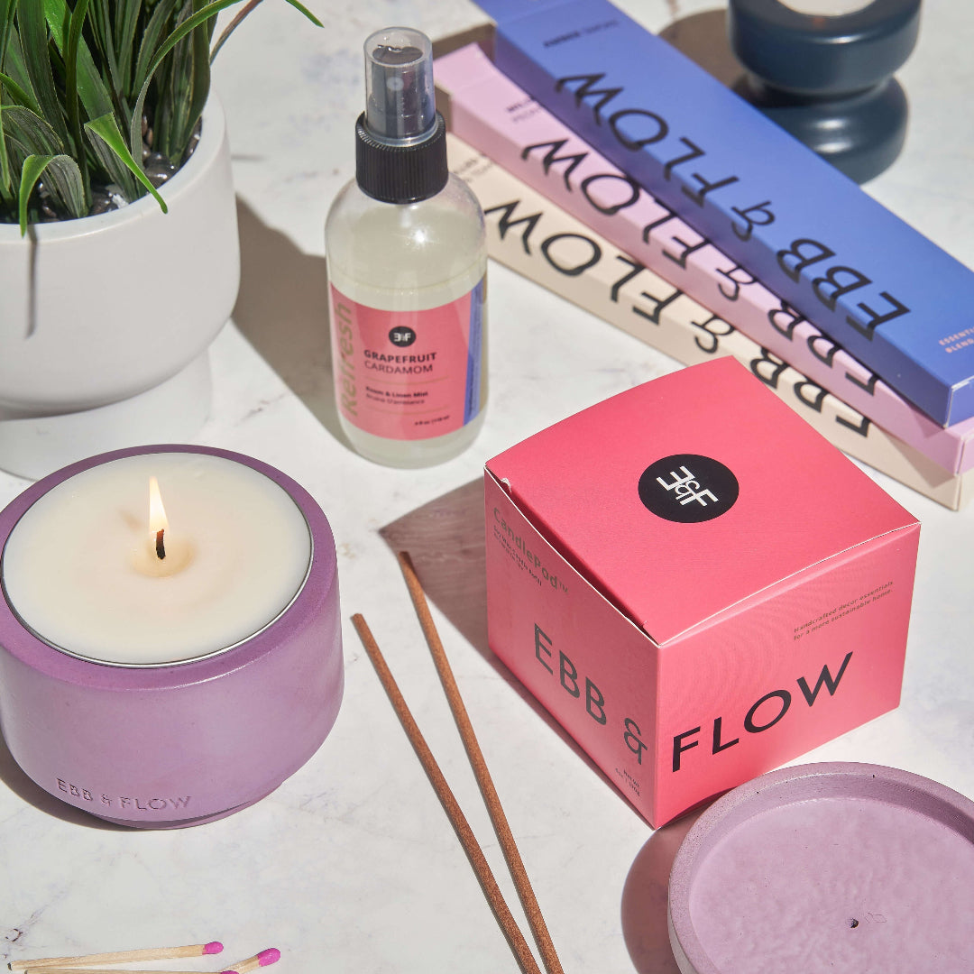 A styled flat-lay of EBB & FLOW home fragrance products on a light marble surface, including a pink candle box, a pink candle jar with a lit candle, a clear spray bottle, incense sticks, matches, a plant in a white pot, and a blue candle holder.