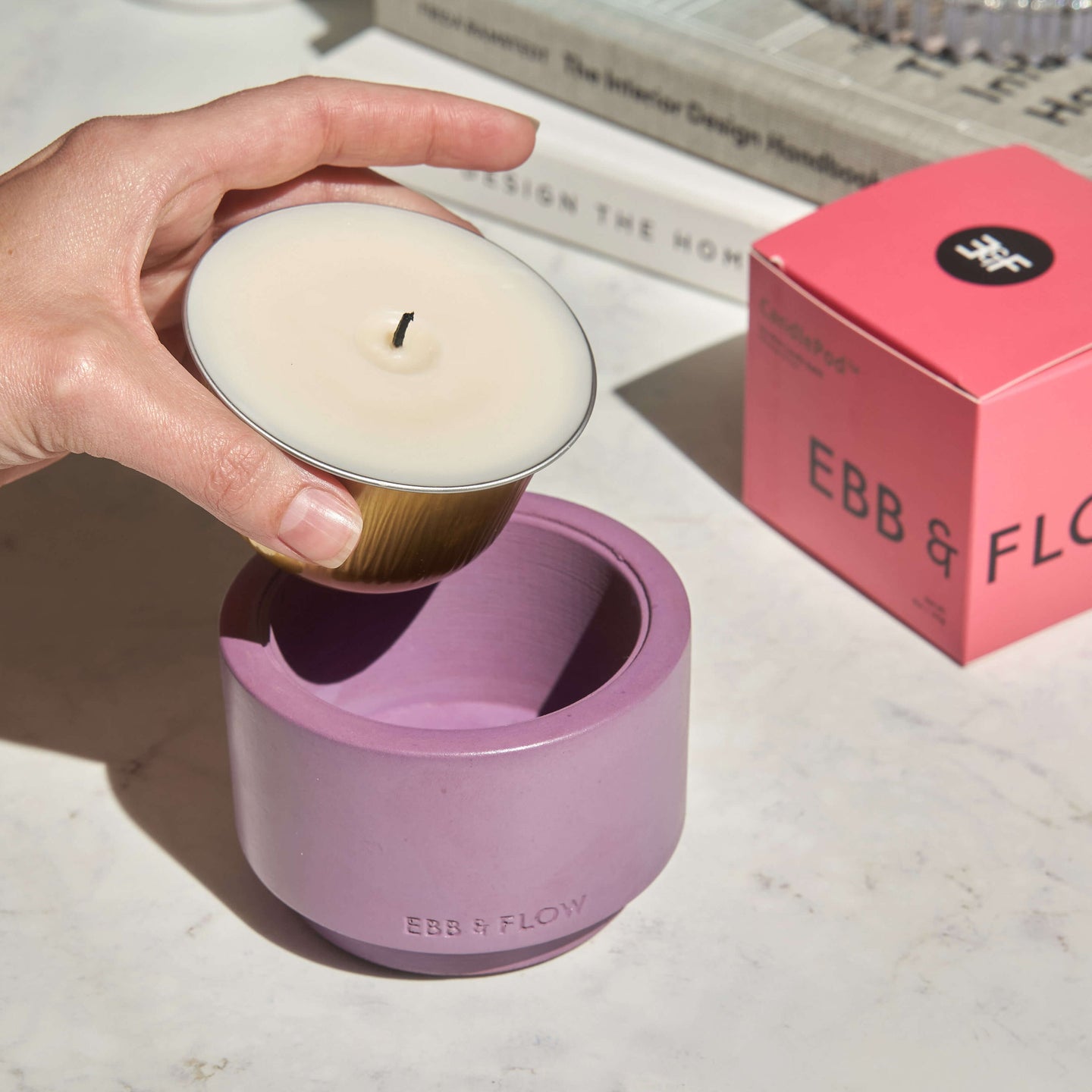 Hand holding a small candle inside a purple container with 'Ebb & Flow' branding, on a surface with books and a pink box.