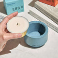 Hand holding a candle with a blue container labeled 'Ebb & Flow' on a surface with books and boxes.