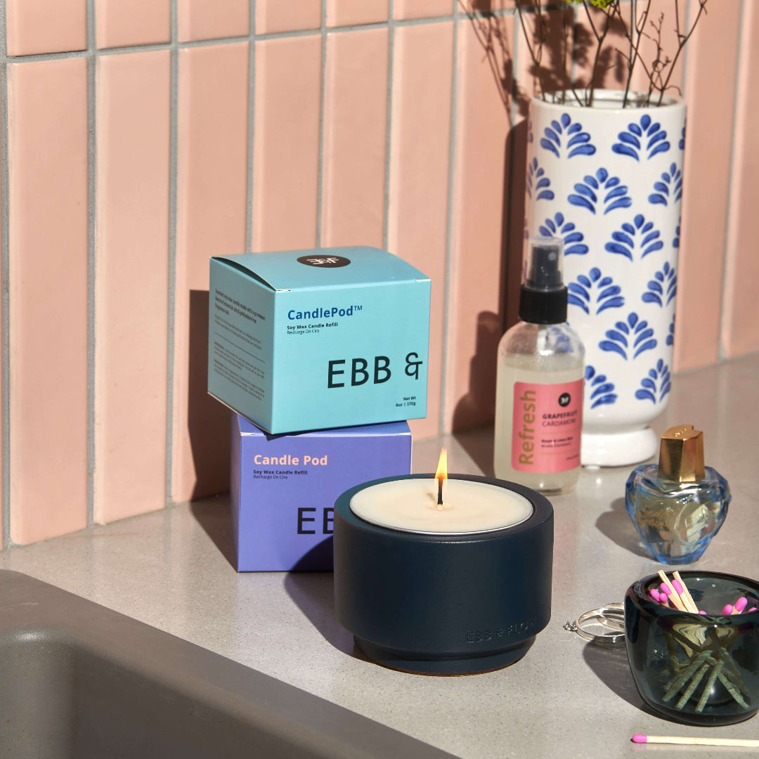 A lit EBB & CandlePod scented candle in a matte navy blue holder sits on a countertop, with its stacked light blue and purple product boxes behind it. A pink-and-white patterned vase with greenery, a small bottle, and a match cup are nearby against a pink tiled wall in bright sunlight.