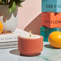 A lit EBB & FLOW candle in a terracotta-colored holder sits on a marble surface, styled with books, a potted plant, oranges, and stacked product boxes in the background.