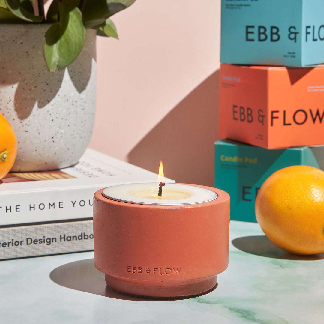 A lit EBB & FLOW candle in a terracotta-colored holder sits on a marble surface, styled with books, a potted plant, oranges, and stacked product boxes in the background.